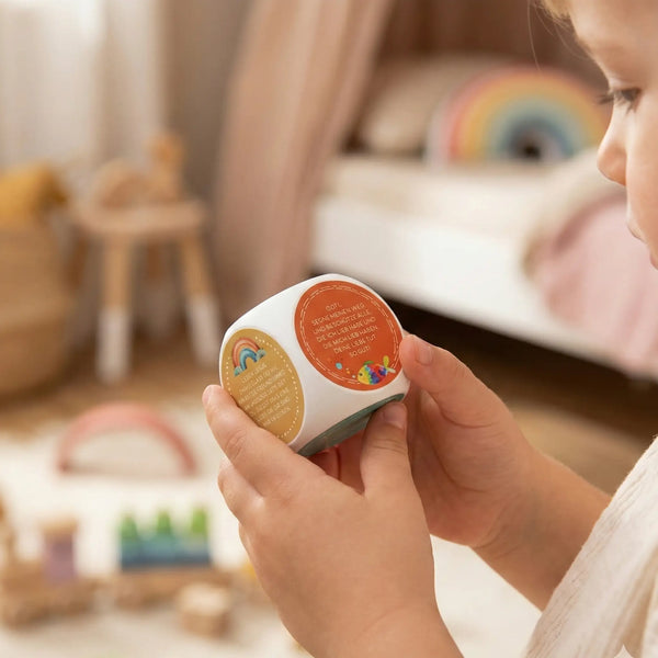Kleinkind hält Gebetswürfel „Dankgebete“ für Kinder – christliches Geschenk mit kurzen Tischgebeten und bunten Motiven, im Hintergrund Kinderzimmer
