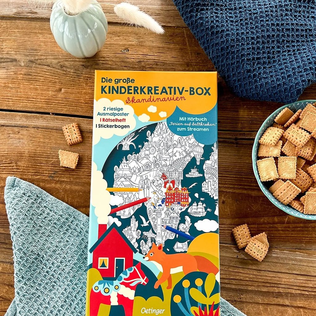 Das Stimmungsbild enthält die Kinderkreativ-Box auf einem Holztisch mit einer Keksschale, einer Trockenblume in einer Vase und blaue Tücher.