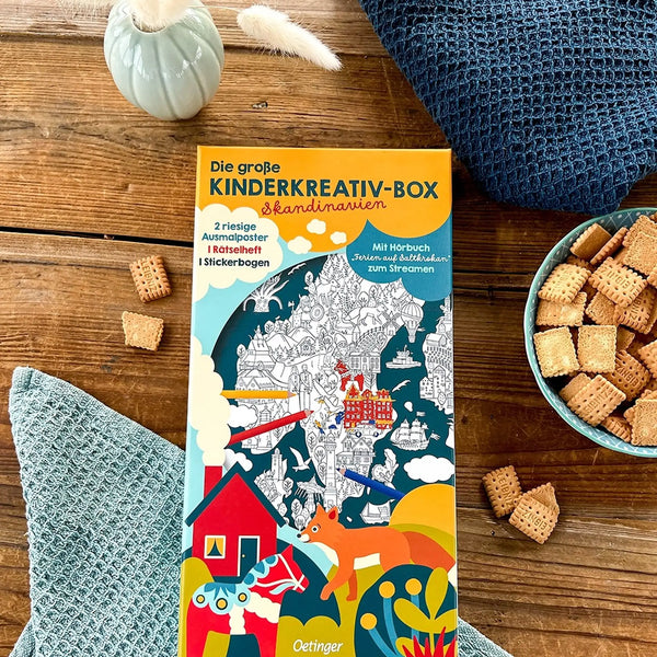 Das Stimmungsbild enthält die Kinderkreativ-Box auf einem Holztisch mit einer Keksschale, einer Trockenblume in einer Vase und blaue Tücher.