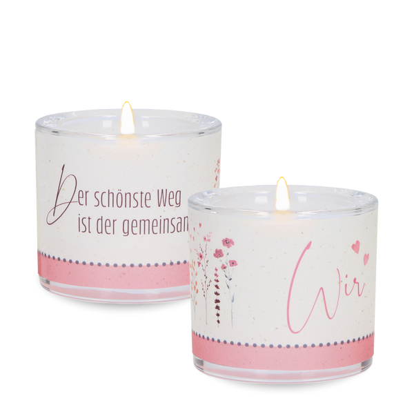 Zwei dekorative weiße Duftkerzen in transparenten Gläsern mit rosa Gestaltung, Blumenmotiven und liebevollen Schriftzügen. Die Kerzen sind angezündet und auf schwarzem Hintergrund dargestellt.