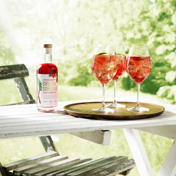 Flasche mit Aperitif, 3 Gläser mit Mixgetränk und Eiswürfeln und Limette auf Tablett, auf Gartentisch, im Hintergrund Gartenszene, grüne Pflanzen, Wiese