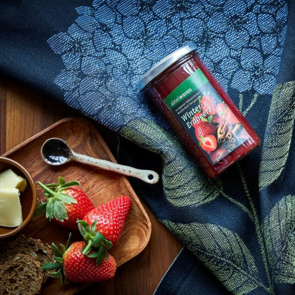 Ein Marmeladenglas liegt auf einem Holztisch mit blauer Tischdecke, rechts liegen Erbeeren und Zimt
